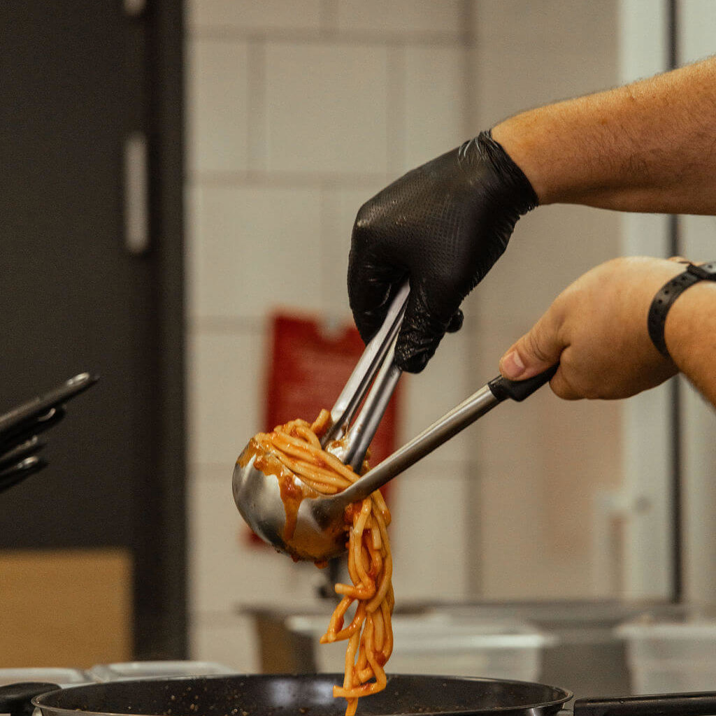 Chef en cuisine manipulant des spaghetti avec des pinces, prêt à servir le plat dans un cadre professionnel et moderne.