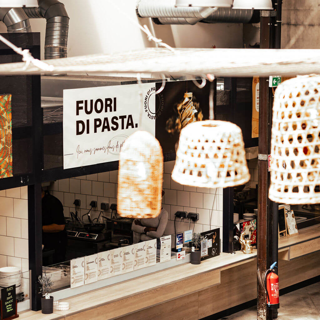 Vue d’un stand de La Commune avec une enseigne « Fuori di Pasta » et des suspensions décoratives en osier, ambiance industrielle.