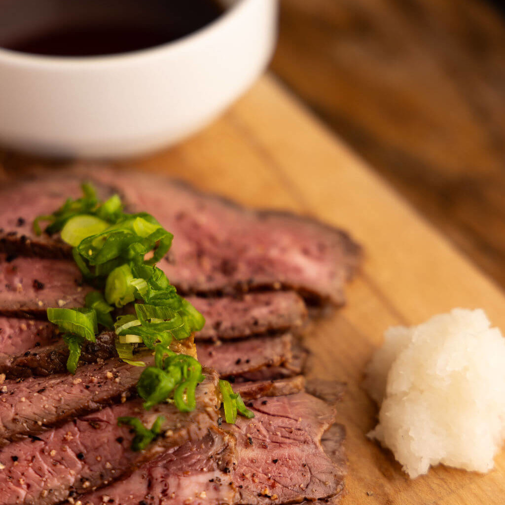 Tranches de bœuf grillé japonais servies sur une planche en bois avec garniture de cébette et accompagnées d'une sauce umami.