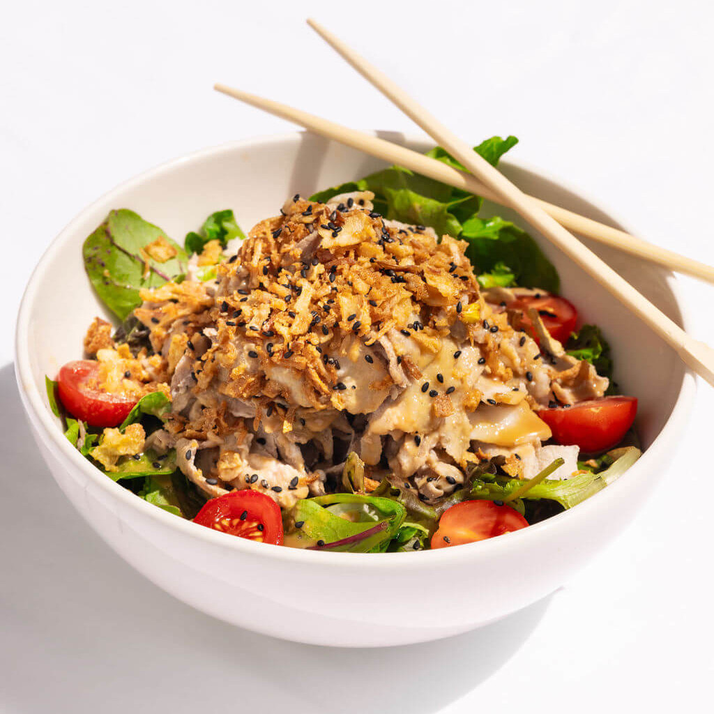 Salade japonaise garnie de nouilles, tomates cerises et oignons frits dans un bol, accompagnée de baguettes en bois.