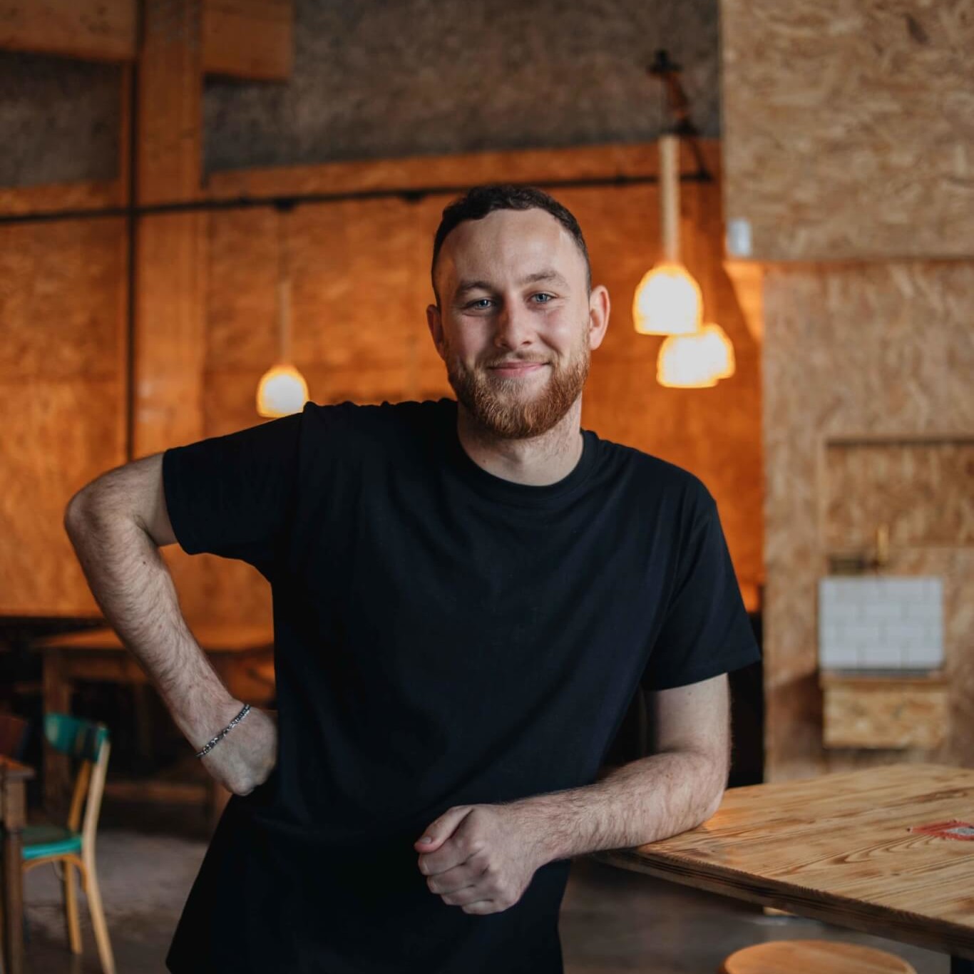 Un chef barbu, bras croisés, souriant dans un espace chaleureux et lumineux du restaurant.