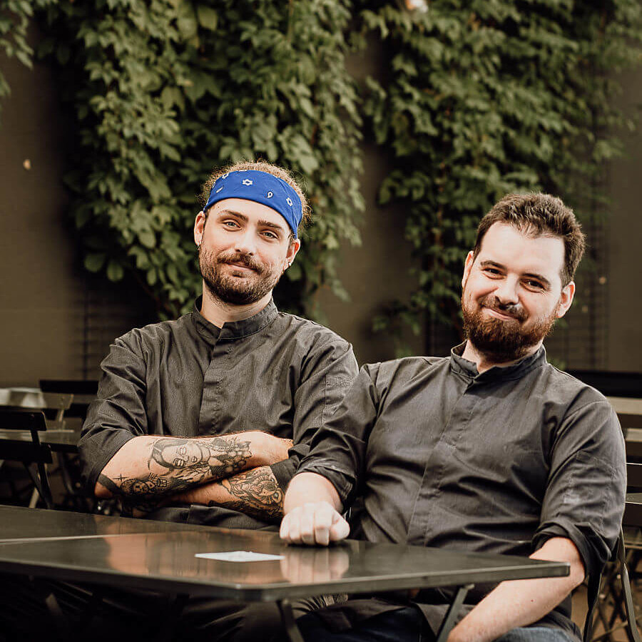 Deux chefs posent avec un sourire, assis à une table extérieure, devant un mur végétal.