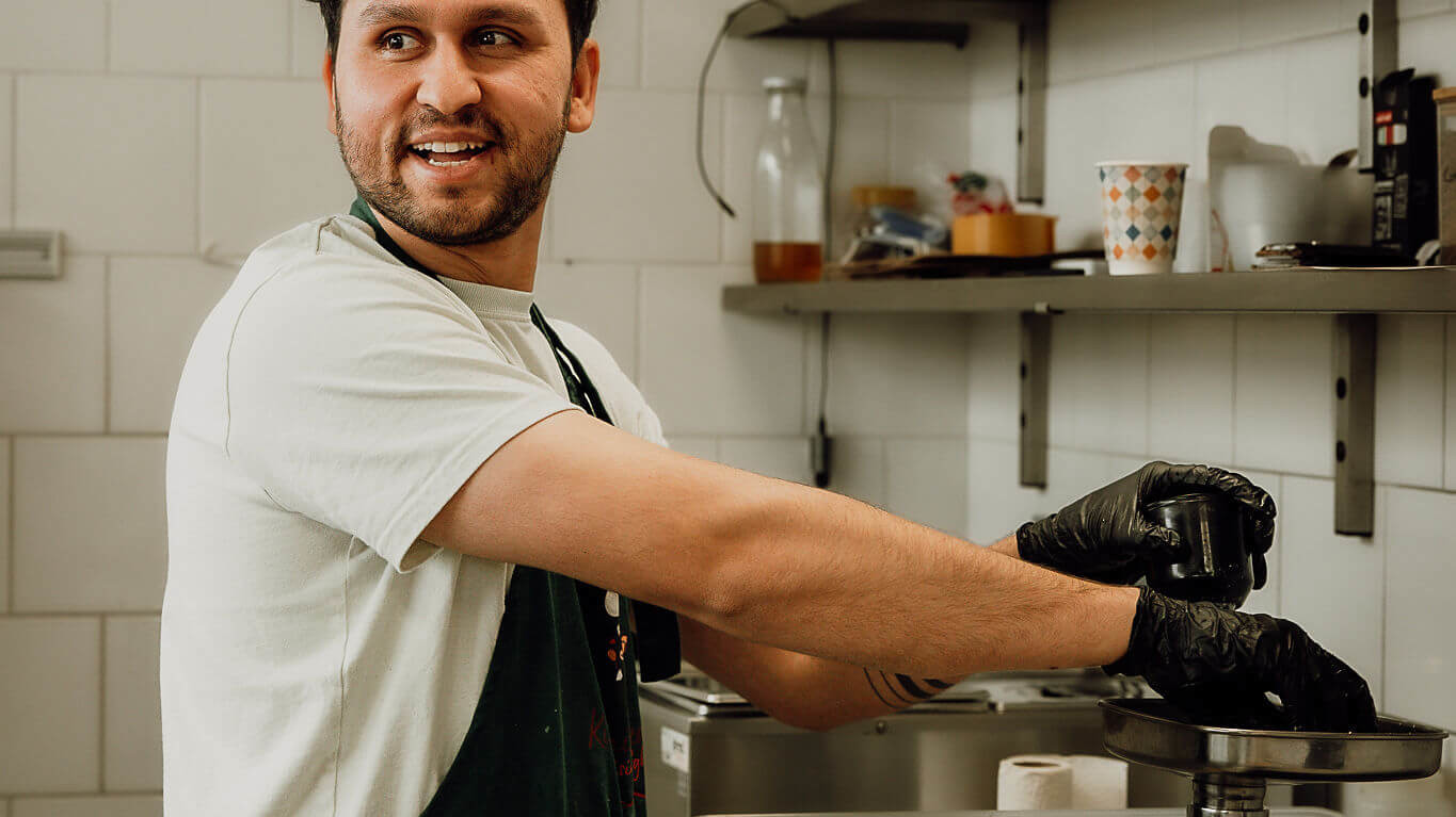 Un chef en cuisine, portant un tablier vert, prépare des ingrédients avec des gants noirs, souriant tout en travaillant dans un environnement dynamique.