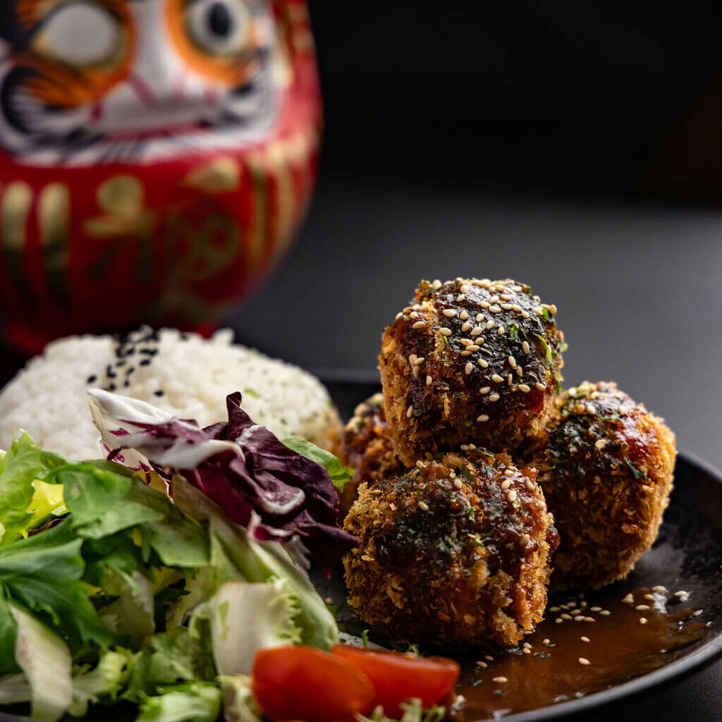 Boulettes japonaises croustillantes accompagnées de riz blanc, de salade verte et de sauce, servies dans une assiette noire avec un daruma rouge en arrière-plan.