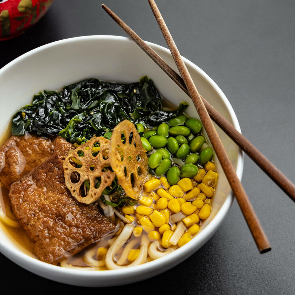 Bol de ramen japonais garni de légumes frais, de maïs, de lotus croustillant et de tofu, avec un daruma rouge décoratif en arrière-plan.