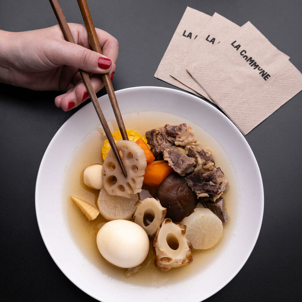 lat asiatique traditionnel composé d'ingrédients variés, tels que du lotus, des œufs et des légumes, présenté dans un bol blanc avec des baguettes en bois, accompagné de serviettes La Commune.