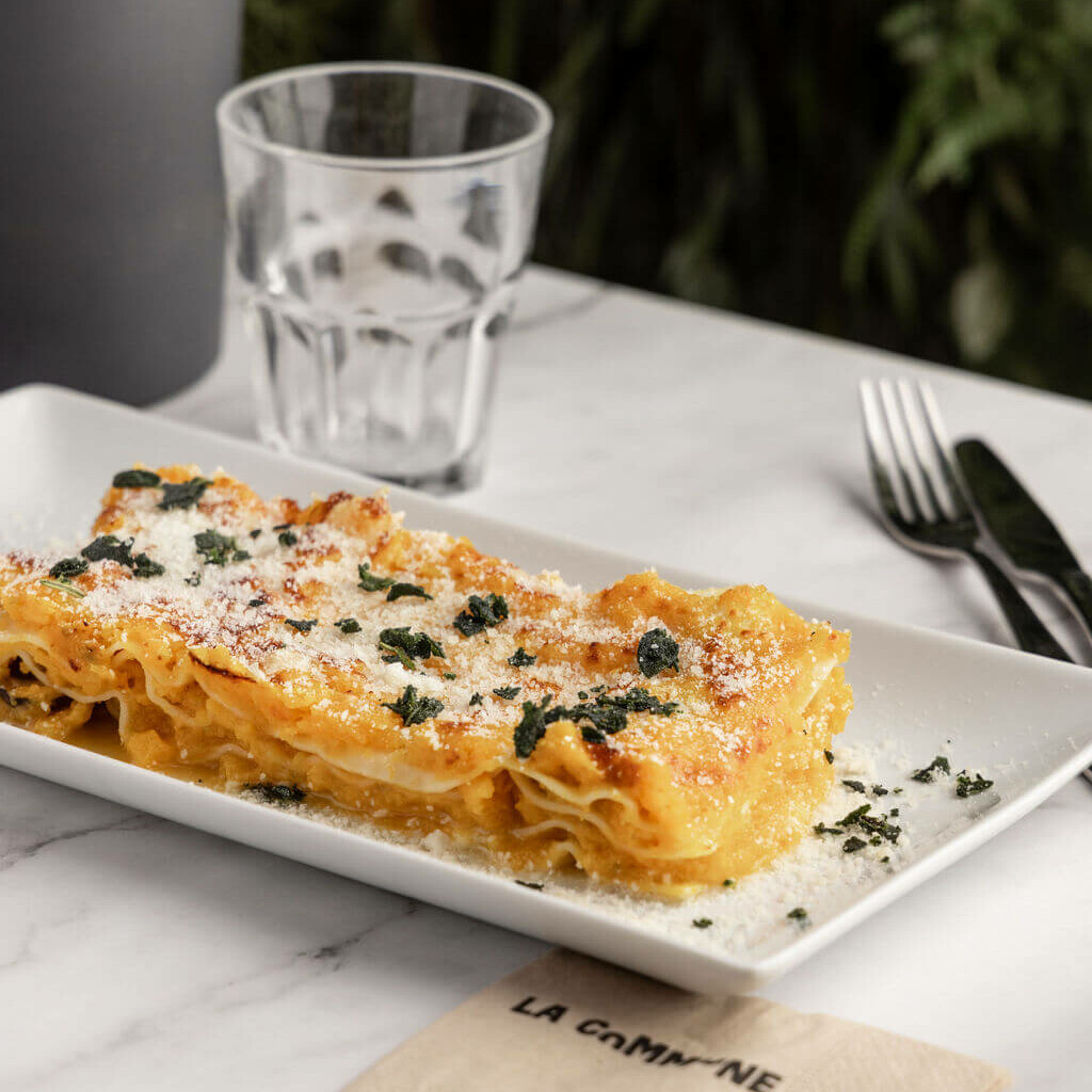 Lasagnes maison à la courge et au fromage, garnies de sauge et de parmesan, présentées sur une assiette blanche avec une serviette La Commune.