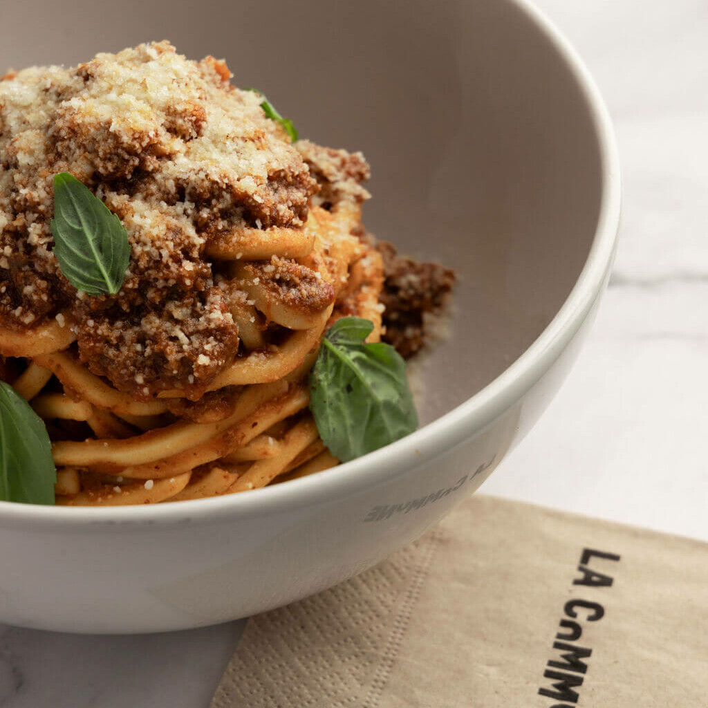 Bol de pâtes à la bolognaise avec parmesan râpé et feuilles de basilic frais, accompagné d'une serviette La Commune sur une table en marbre.