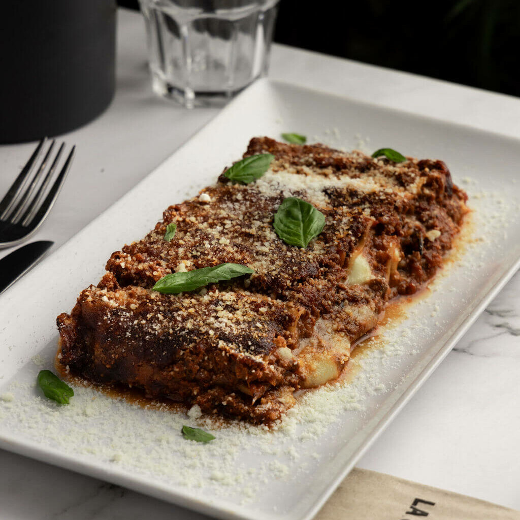 Lasagnes traditionnelles garnies de sauce bolognaise, fromage et basilic, servies sur une assiette blanche avec un verre en arrière-plan.