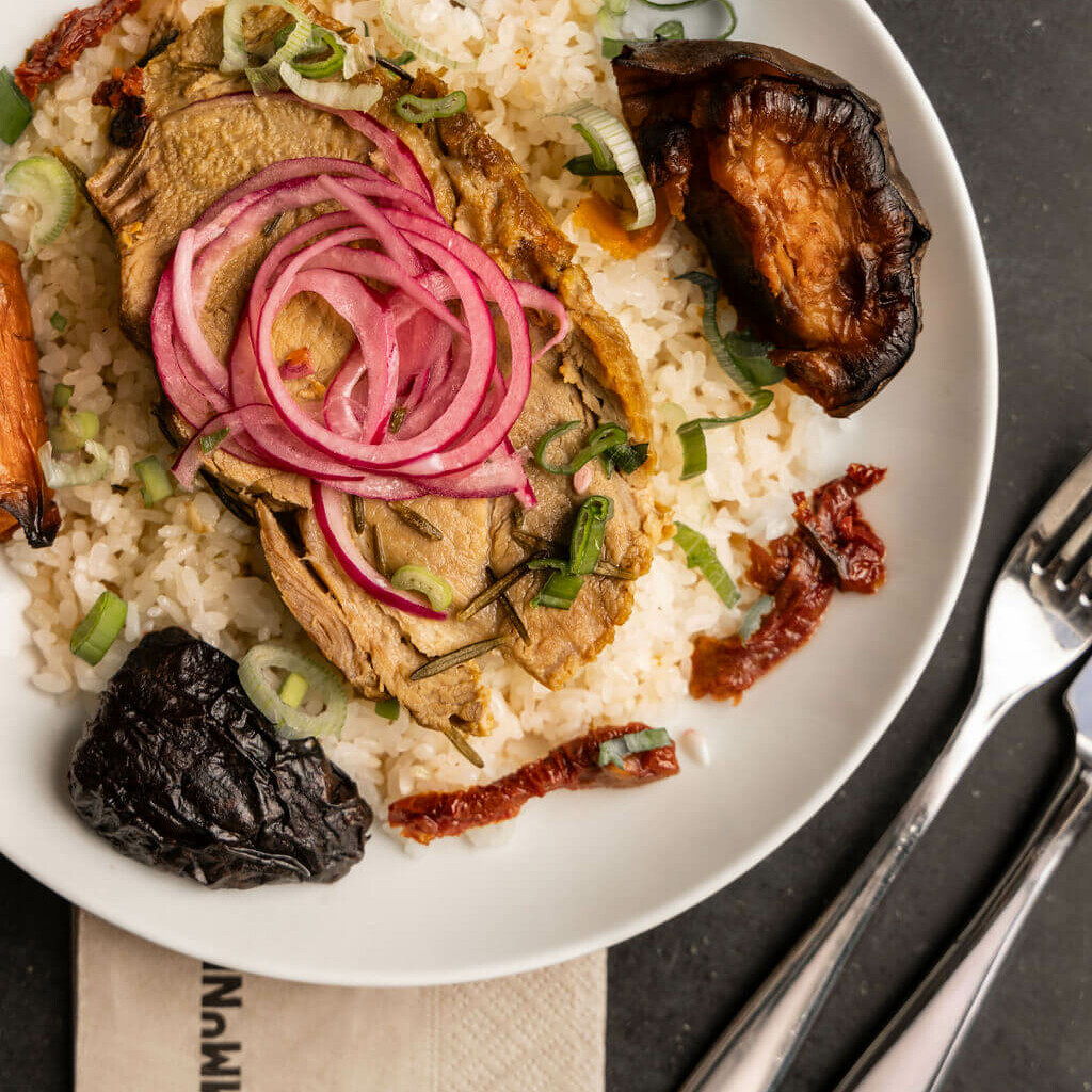 Assiette gastronomique avec une tranche de viande braisée garnie d’oignons rouges marinés, servie sur du riz avec légumes rôtis et tomates séchées.