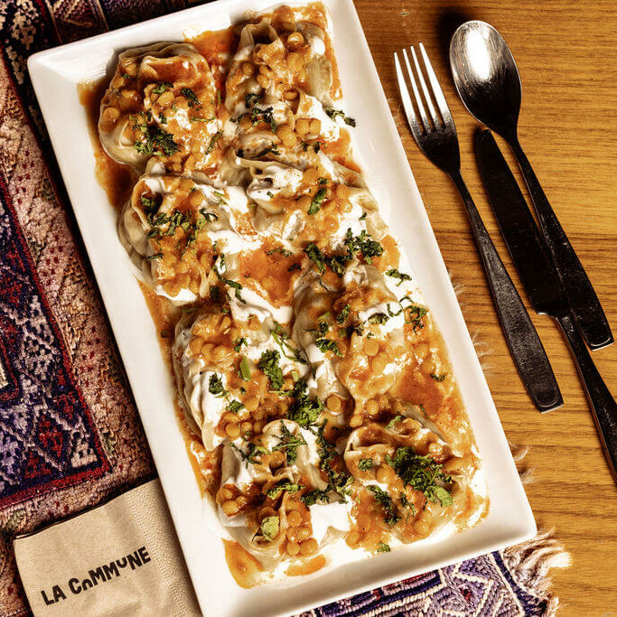 Assiette de mantous afghans nappés de sauce tomate, lentilles et yaourt, servis sur nappe traditionnelle à La Commune.