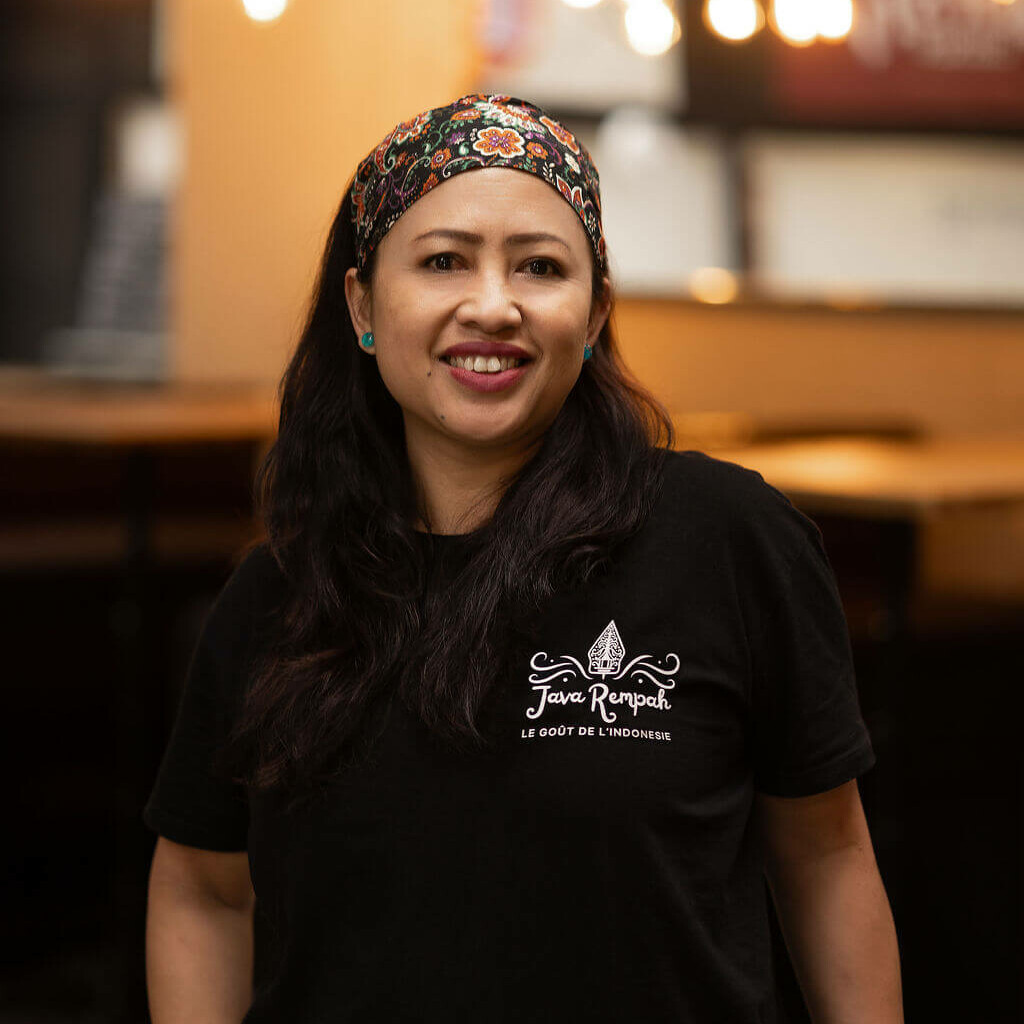 Portrait de la cheffe de Java Rempah portant un bandeau fleuri, souriante dans un décor chaleureux de restaurant indonésien
