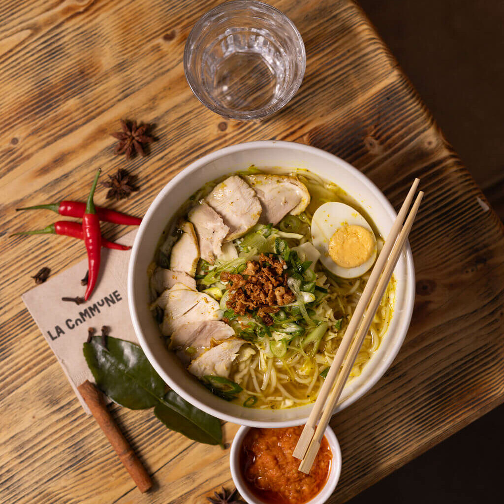 Bol de soupe indonésienne avec poulet, œuf dur, germes de soja et ciboule, servi avec sauce pimentée, baguettes et condiments sur table en bois.