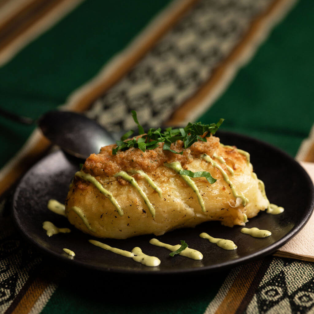 Pomme de terre farcie au poulet et herbes, nappée de sauce verte sur une assiette noire, un incontournable de la cuisine andine.