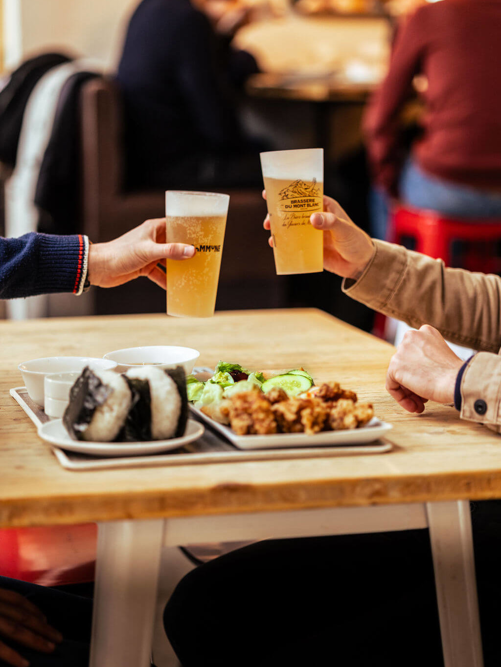 Deux personnes trinquent avec des bières artisanales à La Commune, accompagnées de plats gourmands de street-food asiatique.