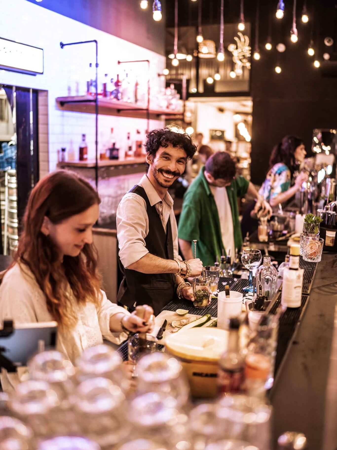 Barman souriant préparant des cocktails artisanaux dans l’ambiance festive et chaleureuse de La Commune à Lyon.