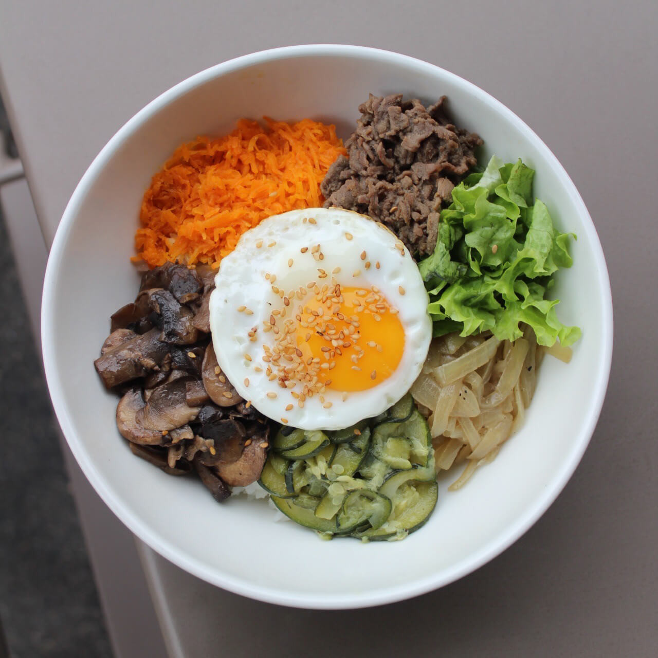 Bol de Bibimbap coréen avec œuf au plat, carottes râpées, champignons, bœuf mariné, salade verte et riz, garni de graines de sésame.