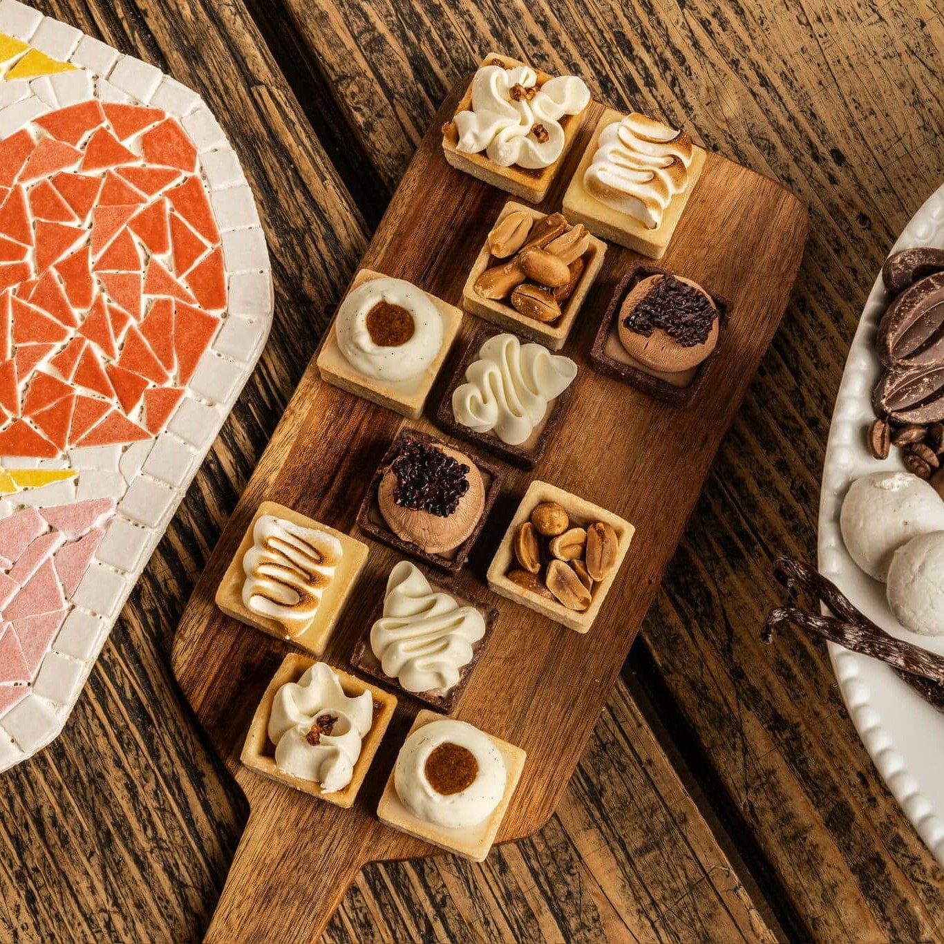 Assortiment de mini-tartelettes gourmandes sur une planche en bois, accompagné de mosaïque colorée et d'une assiette avec citrons, noix, chocolat et vanille.