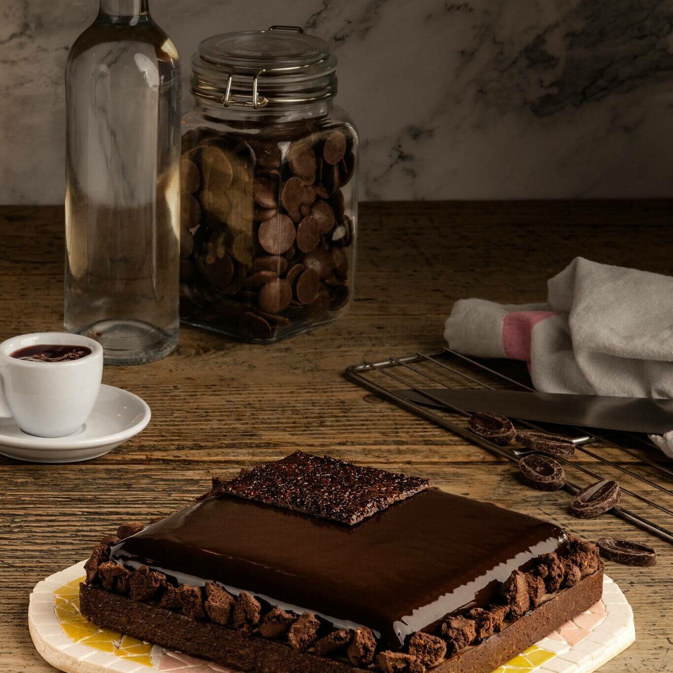 Pâtisserie au chocolat brillant présentée avec une tasse de café, une bouteille et un bocal de pépites de chocolat, sur une table en bois.