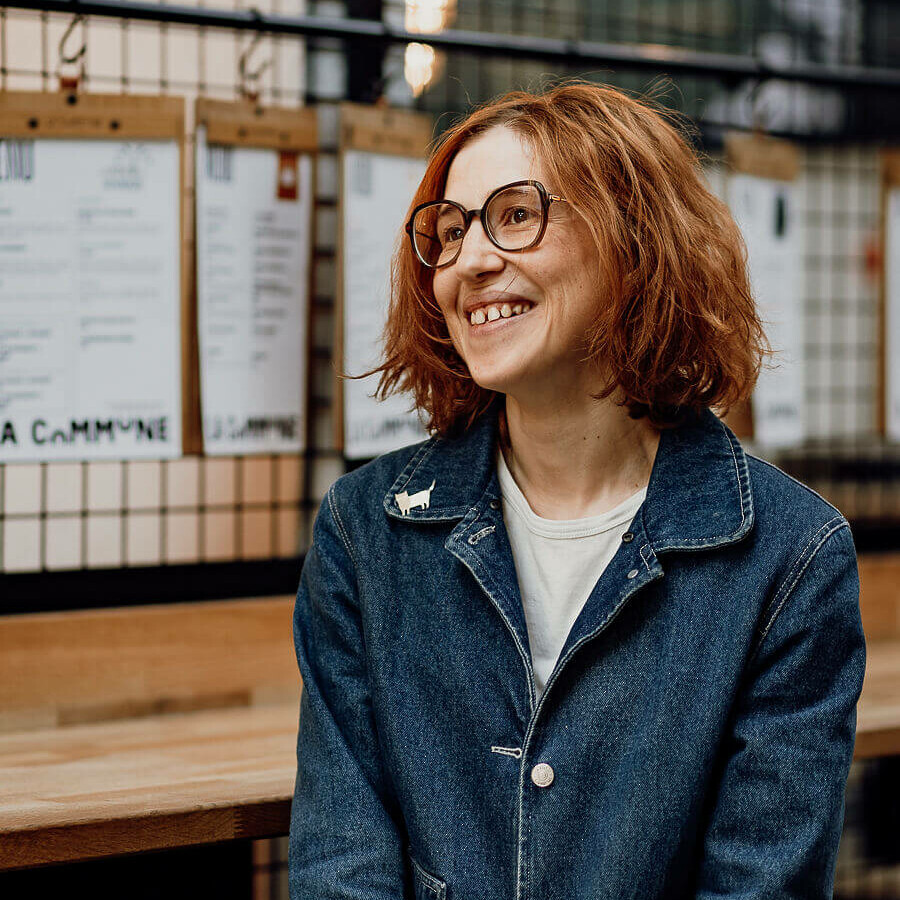 Portrait d'une femme souriante avec des cheveux roux et des lunettes, portant une veste en jean, posant dans un espace chaleureux à La Commune.