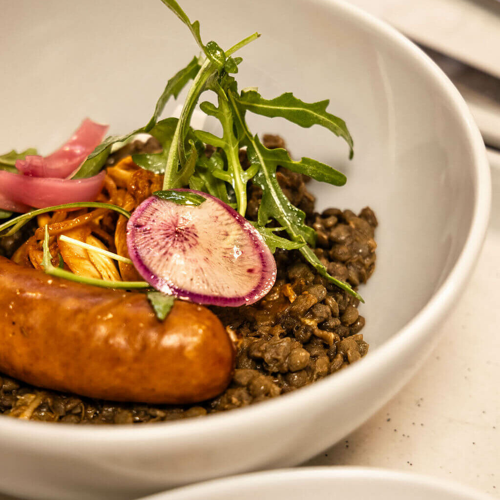 : Assiette de lentilles avec saucisse, légumes marinés et roquette, servie dans un bol blanc.