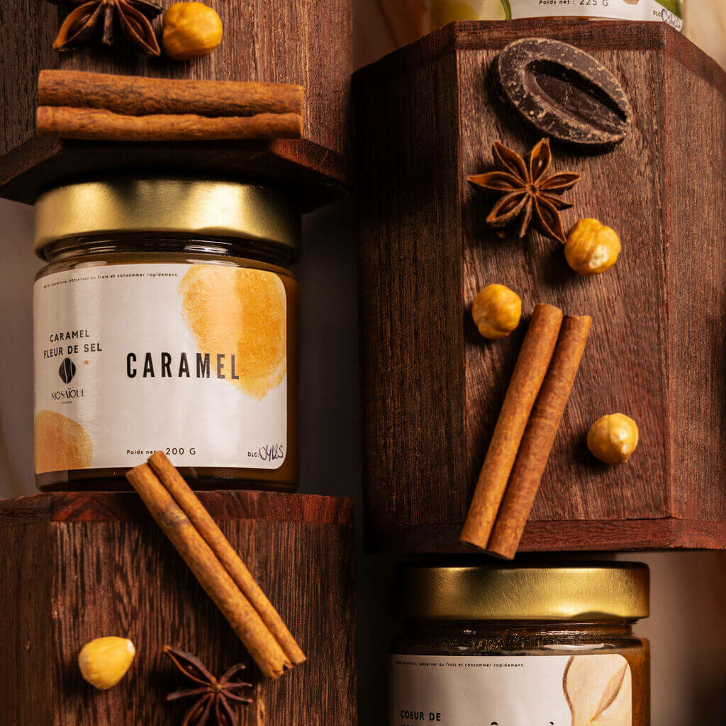Pots de caramel à la fleur de sel et pâte à tartiner noisette Mos Voile, accompagnés d’épices et de chocolat, disposés sur des blocs en bois