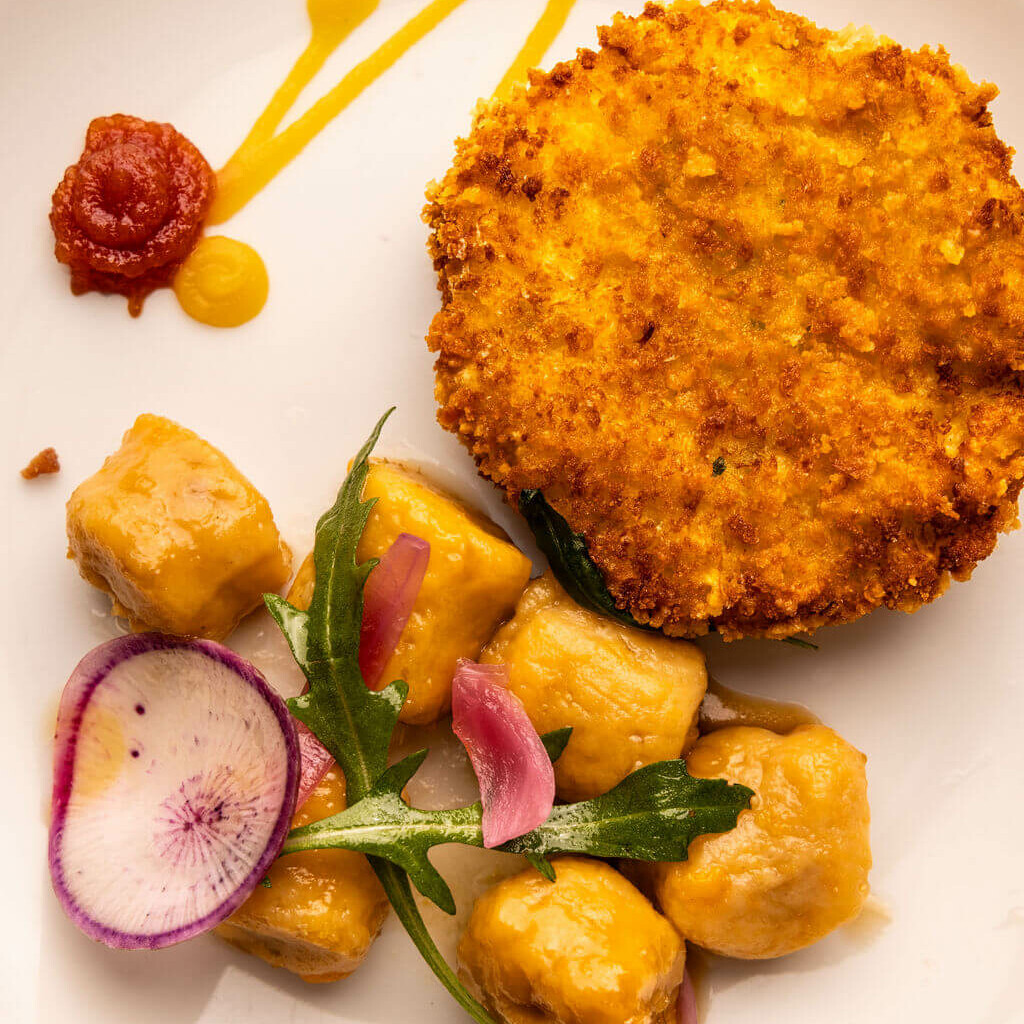 Assiette de gnocchis accompagnés d’une galette panée croustillante, avec sauce et légumes colorés.